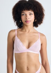 Vaaleanpunainen bralette, jossa on pitsireunus kauluksessa, säädettävät olkaimet ja sileä kangasrunko. Minimalistinen muotoilu ilman pehmusteita.