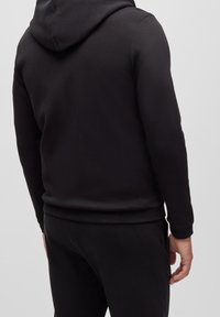 En person som bär en svart huvtröja och matchande svarta byxor, sedd bakifrån mot en enkel ljus bakgrund.