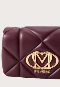 Bordeauxleren clutch met een geborduurd ontwerp, voorzien van een gouden logo-accent. Glad oppervlak met kruisende lijnen en afgeronde hoeken.