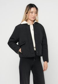 Veste matelassée noire avec une silhouette structurée, dotée d'un col montant, de boutons sur le devant et de poches latérales ; portée sur un pull crème.
