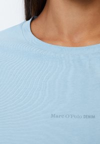 Light blue cotton t-shirt with a crew neckline. Features a smooth texture and includes a subtle logo in the lower right corner.