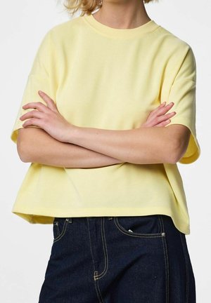 Persoon draagt een lichtgeel T-shirt met korte mouwen met gekruiste armen en donkerblauwe spijkerbroek met zichtbare gele stiksels.