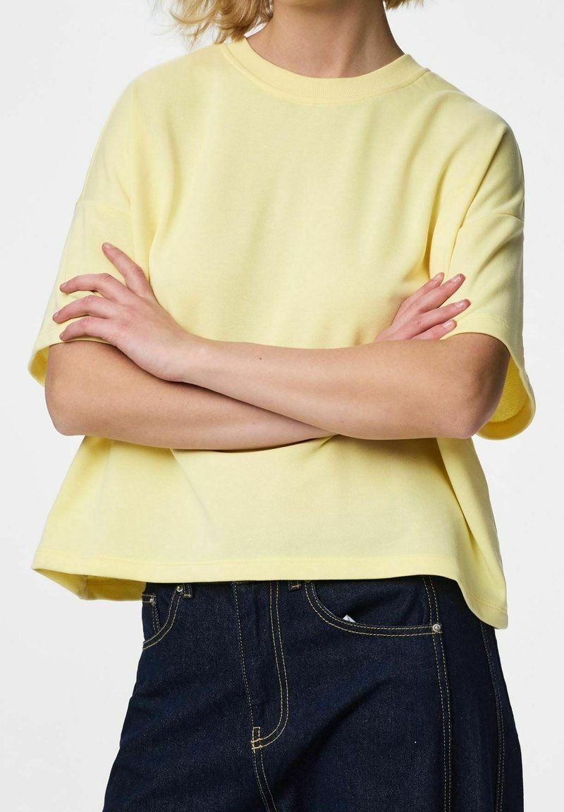 Personne portant un haut jaune clair à manches courtes, bras croisés, et un jean bleu foncé avec des coutures jaunes visibles.