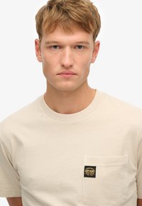 T-shirt beige a maniche corte realizzata in cotone, con taschino sul petto dotato di un patch logo nero e giallo. Collo a giro e design semplice.
