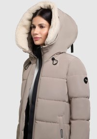 Jachetă puffer bej cu o glugă mare căptușită cu blană artificială. Dispune de un fermoar frontal, accente din metal negru și o etichetă branduită pe mânecă.