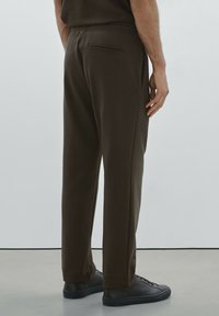 Braune, lässig geschnittene Jogginghosen mit elastischem Bund, Gesäßtasche und schmal zulaufenden Beinen. Getragen mit schwarzen, strukturierten Sneakers. Glatte Stofftextur.