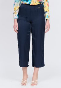 Pantaloni accorciati blu navy con una texture liscia, elastico in vita e aperture laterali all'orlo, abbinati a sandali bianchi con cinturino.