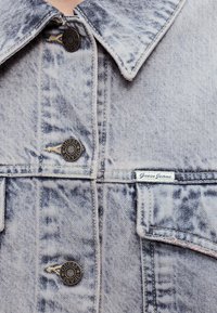 Φωτεινό γαλάζιο τζιν μπουφάν με κόλλημα οξέος και μεταλλικά κουμπιά με την ετικέτα "Guess Jeans" και μια μικρή λευκή υφασμάτινη ετικέτα με το κείμενο "Guess Jeans" κοντά στην τσέπη στο στήθος.