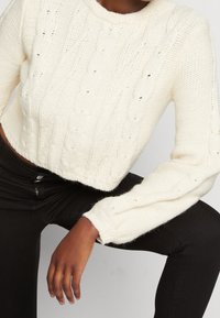 Vit stickad kort tröja med kabelmönster, rund hals och långa ärmar. Bärs med svarta tajta jeans. Strukturerad tyg.