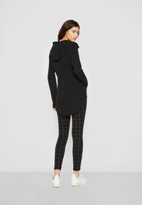 Haut à capuche noir avec un dos long, associé à des leggings noirs avec un motif à carreaux blancs, et des baskets blanches. Texture de tissu lisse.