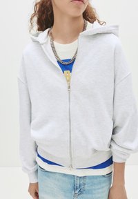 Lys grå zip-up hættetrøje med en afslappet pasform, ribbede manchetter og talje. Lagd over en blå t-shirt med gyldne bogstaver. Sølv kæder er synlige.