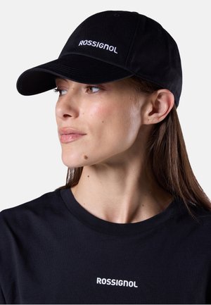 Vrouw met een zwarte Rossignol-pet en bijpassend zwart Rossignol-shirt, kijkt iets naar links tegen een effen achtergrond.