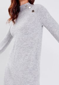 Robe pull tricotée gris clair avec manches longues, texture côtelée, col montant et boutons décoratifs sur l'épaule.