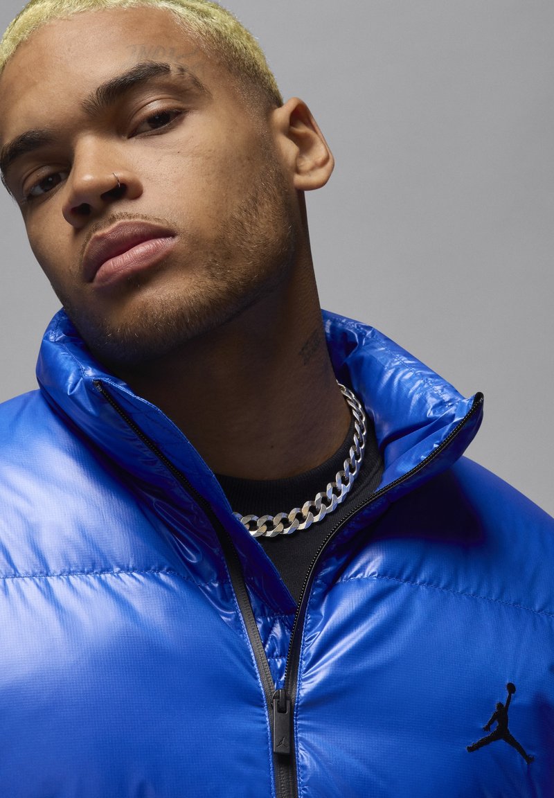 Chaqueta de plumas azul con textura brillante, cierre con cremallera y logo de Jordan en negro. Lleva puesta una camiseta negra y está accessorized con un collar de cadena de plata.