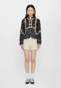 Blusa nera a fiori con maniche lunghe, dettagli crema e scollo alto, abbinata a pantaloni corti beige e scarpe nere con cinturini.