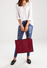Borsa tote in pelle bordeaux con dettagli neri, due manici e apertura superiore. Indossata con una camicia bianca con pois e jeans blu strappati.
