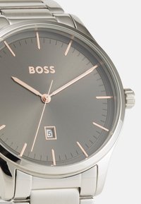 Reloj de acero inoxidable plateado con una esfera gris, acentos en oro rosa, marcadores de hora minimalistas y una función de fecha en la posición de las 6 en punto.