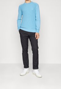 Helles blaues langärmliges geripptes Shirt mit Rundhalsausschnitt und kleinem Logo, kombiniert mit schwarzen Slim-Fit-Hosen und weißen Sneakers.