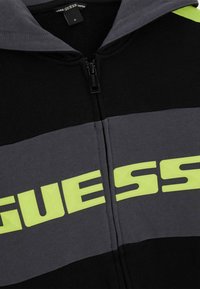 Black and gray zip-up hoodie with a neon yellow "GUESS" logo. Features a smooth texture and a hood with a two-tone design.