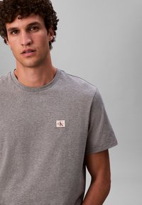 Siva pamučna majica kratkih rukava s okruglim izrezom, s malom CK oznakom na lijevoj strani prsa. Mekana tekstura.