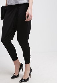 Czarne spodnie haremowe wykonane z miękkiej, elastycznej tkaniny; dopasowane u kostek, o luźnym, drapowanym kroju, zestawione z czarnymi, spiczastymi szpilkami.