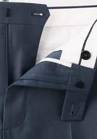 Gros plan de la ceinture d’un pantalon bleu avec bouton, boutonnière, fermeture éclair et doublure intérieure en tissu blanc avec bouton noir et détail de bordure.