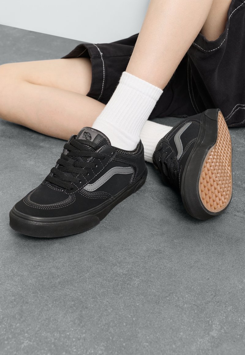 Personne portant des baskets noires avec des chaussettes blanches et un short noir assise sur un sol gris, un pied montrant le motif de la semelle de la chaussure.