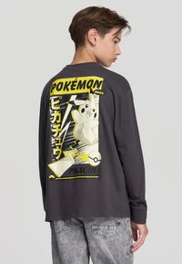 Koszulka z długim rękawem w ciemnoszarym kolorze z żywym motywem Pikachu na plecach, z żółtymi i czarnymi akcentami. Wykonana z materiału bawełnianego, o luźnym kroju.