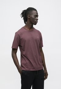 Mladi moški z dreadlocki, oblečen v bordo majico s kratkimi rokavi in črne hlače, stoji z eno roko v žepu in gleda vstran.