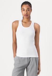 Weißes, geripptes Tanktop mit dünnen Trägern und rundem Ausschnitt, kombiniert mit grauen, plissierten Hosen. Der Stoff hat eine weiche, glatte Textur.
