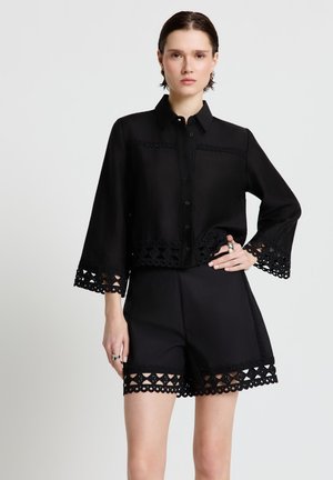 Frau trägt ein schwarzes, geknöpftes Hemd mit Spitzenbesatz an Ärmeln und Saum, kombiniert mit passenden schwarzen Shorts mit Spitzen-Details.