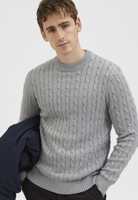 Suéter gris de punto con diseño de trenzas, cuello redondo y puños y dobladillo acanalados. El modelo sostiene una chaqueta azul marino en un brazo.