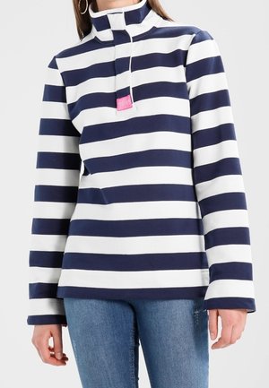 Kvinde iført en marineblå og hvid stribet langærmet pullover med høj krave og halv-lynlås, kombineret med blå jeans.