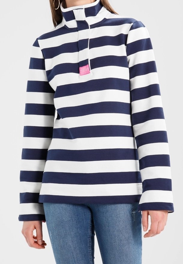 Femme portant un pull à manches longues rayé bleu marine et blanc avec un col montant et une fermeture éclair partielle, associé à un jean bleu.