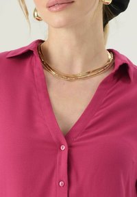 Blusa fucsia con scollatura a V e maniche corte, caratterizzata da chiusure a bottone e una trama liscia. Una collana multi-strato dorata accentua il décolleté.