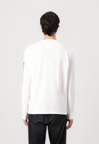 Witte blouse met lange mouwen en een v-vormige halslijn en manchet, gemaakt van gladde stof, met een zwart tekstpatroon langs de linkermouw.