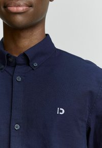 Primer plano de una persona que lleva una camisa de botones azul marino con un pequeño logo bordado en blanco en el pecho.