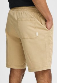 Beige shorts with an elastic waistband and side pockets; made from a smooth fabric with a single back pocket featuring a label.