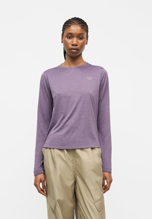 Femme aux cheveux tressés portant un haut de sport à manches longues violet et un pantalon cargo beige, debout devant un fond clair uni.