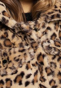 Leopardenmuster Kunstfellmantel mit einer weichen, strukturierten Oberfläche, ausgestattet mit schwarzen Knöpfen und einer Kapuze. Warme Beige- und Brauntöne dominieren das Design.