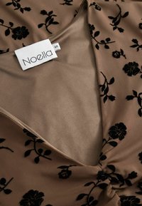 Hnedá textilná blúzka s čiernym kvetinovým vzorom, obsahujúca výstrih do V a zhrnuté detaily v strede. Nálepka označuje "Noella," veľkosť S.