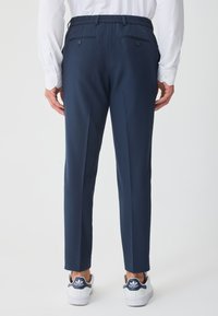 Πίσω όψη ατόμου που φοράει σκούρο μπλε παντελόνι ραμμένο στα μέτρα με κουμπωμένες πίσω τσέπες, λευκό πουκάμισο μακρυμάνικο και λευκά παπούτσια Adidas Stan Smith.