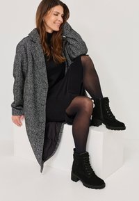 Black suede ankle boots with a block heel and lace-up detail, paired with a herringbone patterned gray oversized coat and a black dress.