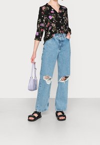Camicia nera con motivo floreale, jeans wide-leg strappati blu, sandali neri e una piccola borsa viola. Materiali strutturati e design informale.