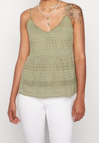Světle zelený krajkový top bez rukávů s výstřihem do V a nastavitelnými ramínky; má horizontální vzory a volný, splývavý design.