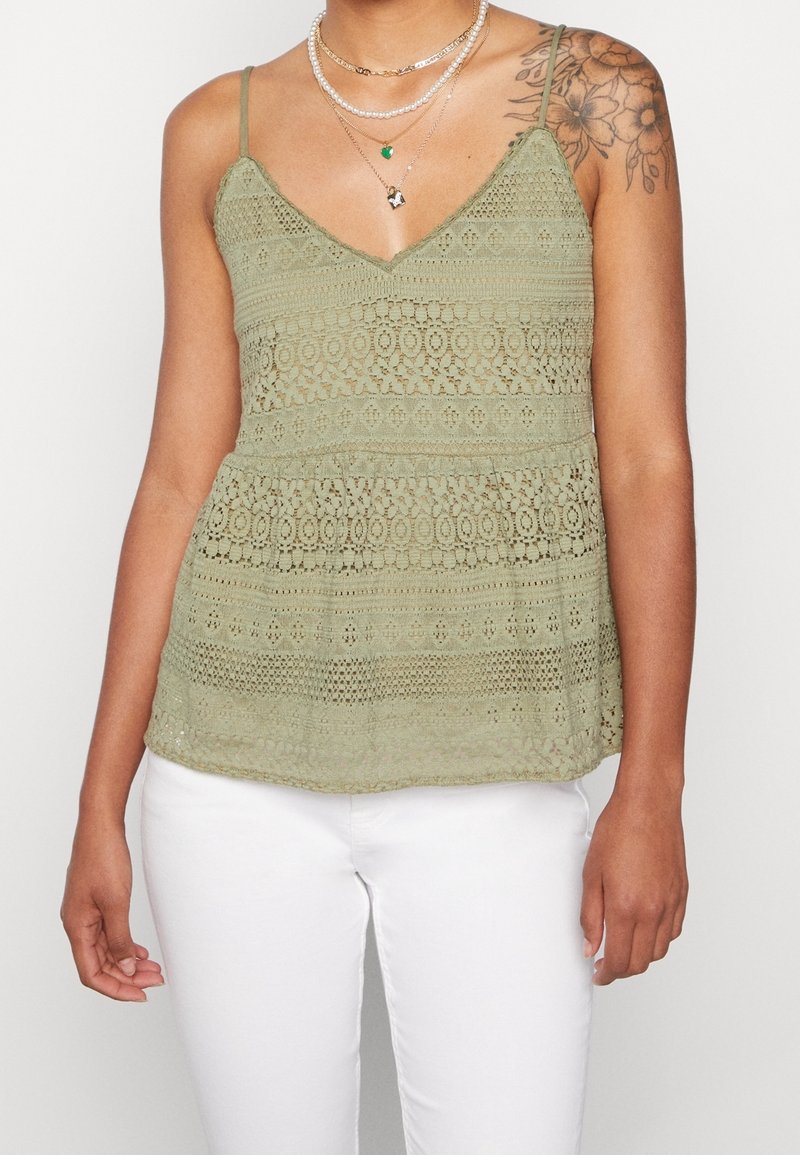 Světle zelený krajkový top bez rukávů s výstřihem do V a nastavitelnými ramínky; má horizontální vzory a volný, splývavý design.