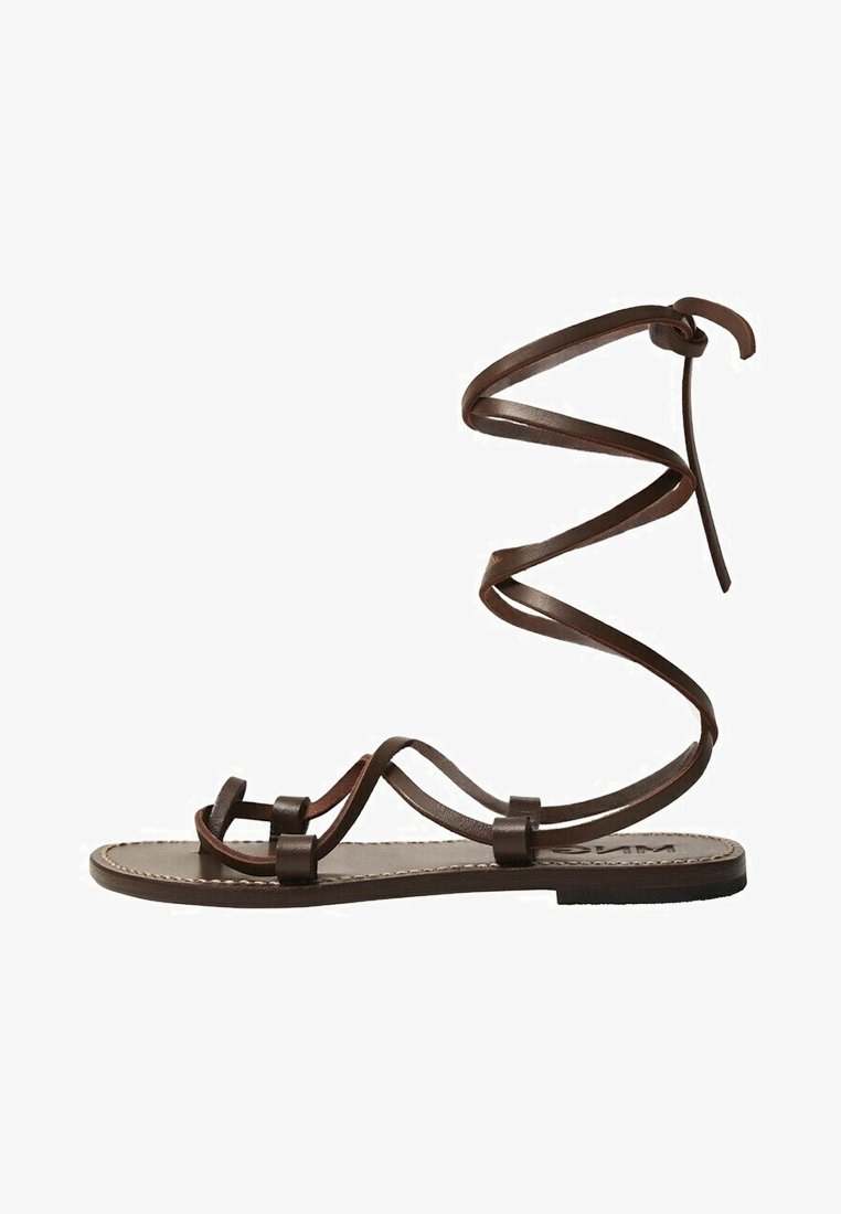 Sandale plate en cuir marron avec des fines lanières et un long lacet qui s'enroule autour de la cheville pour maintenir le pied.