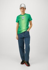 Zaļš gradienta T-kreklis ar vertikālām svītrām, Nike logo un Anglijas emblēmu. Apvienots ar ziliem džinsiem un melniem sneakers ar sarkaniem zoliem.