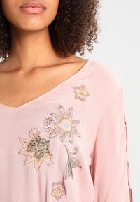 Blusa rosa con scollo a V caratterizzata da ricami floreali in giallo, arancione e verde. Tessuto morbido con un aspetto testurizzato.