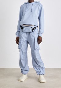 Personne portant un sweat à capuche bleu clair, un pantalon cargo assorti, des chaussures blanches sans lacets et un sac banane bleu clair avec des sangles noires.